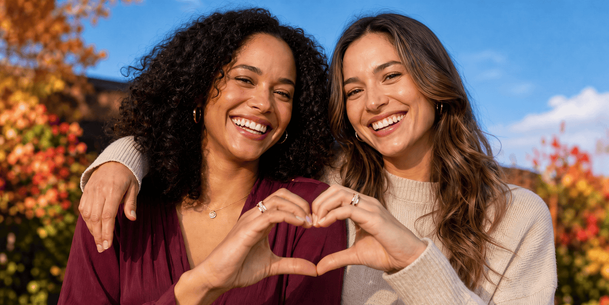 Two women smiling and posing outdoors, forming a heart shape with their hands wearing their stylish smart rings. The woman on the left wears a deep burgundy top, and the woman on the right wears a cream knit sweater.