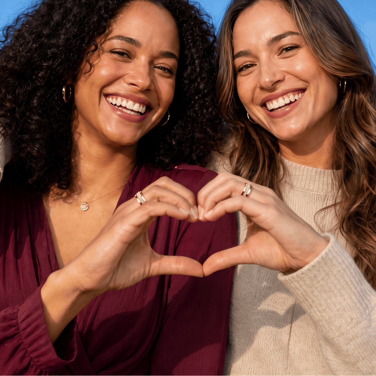 Two women smiling and posing outdoors, forming a heart shape with their hands wearing nala smart rings. The woman on the left wears a deep burgundy top, and the woman on the right wears a cream knit sweater.