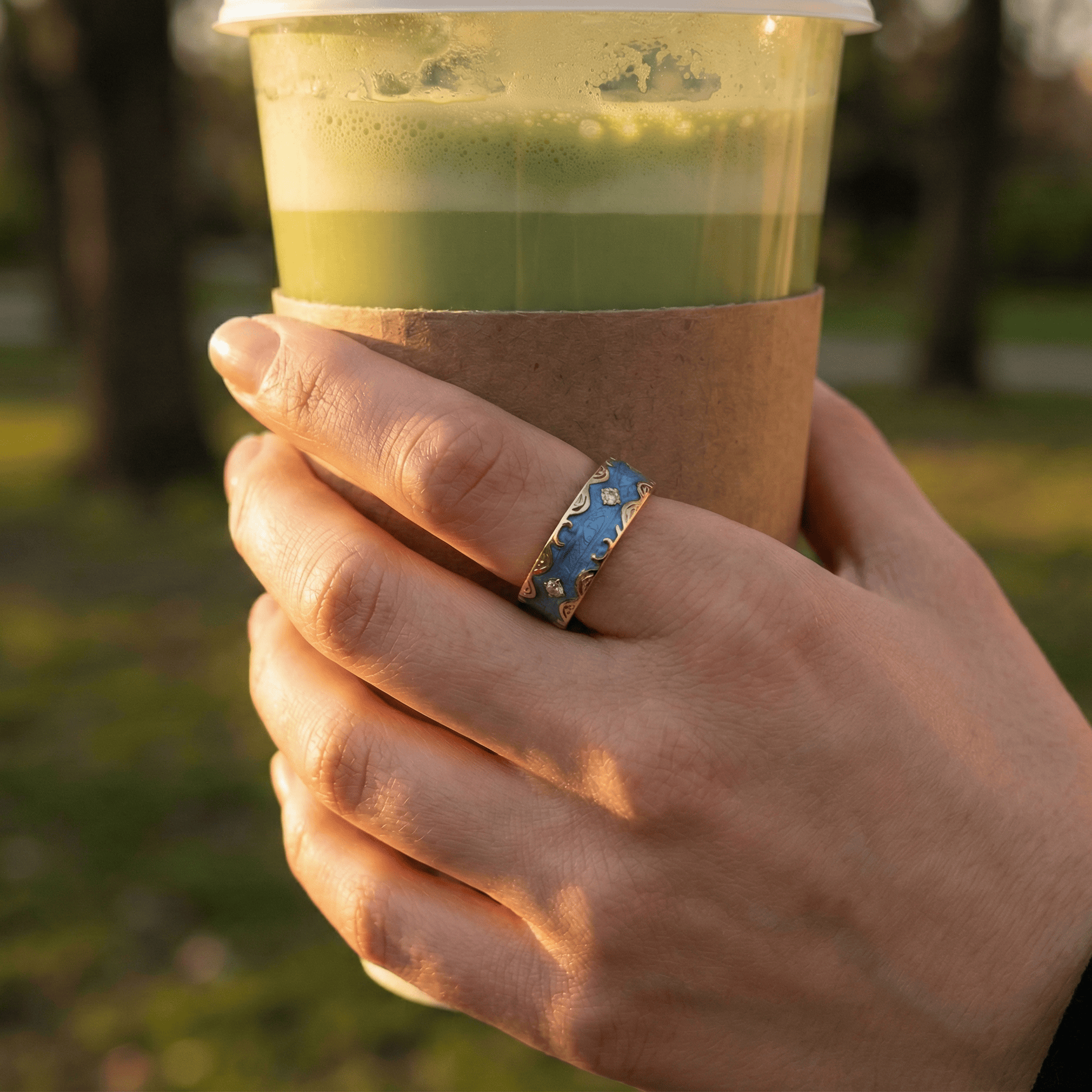 Hand wearing a nala stylish smart ring holding a layered drink in a clear cup with a blurred outdoor background