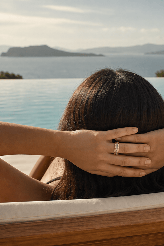 A woman relaxes in a wooden lounge chair by an infinity pool, seen from behind with her hands behind her head. She is wearing a gold ring on one finger, with a scenic ocean and island landscape in the background. nala sells fashionable smart rings.