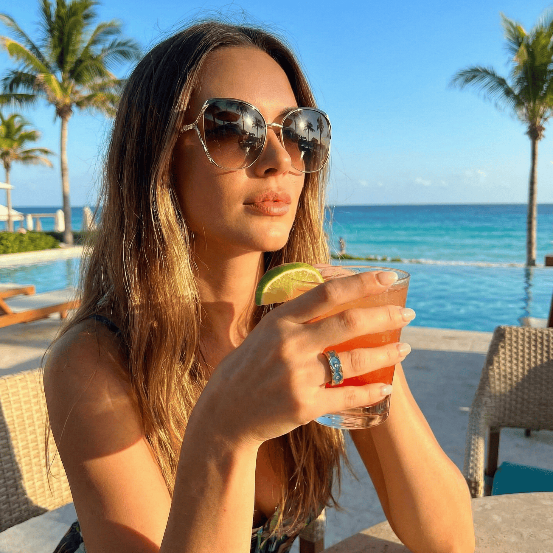 Woman enjoying a cocktail by a pool with palm trees and ocean view, wearing fashion smart ring